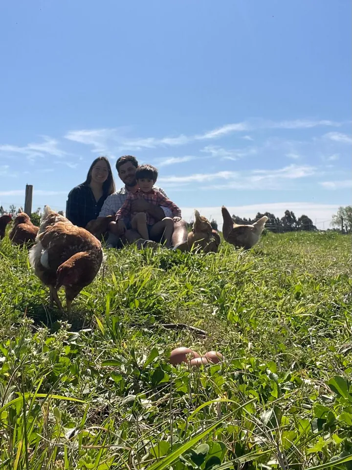 Andrés, Nadia y Felipe sentados en el pasto, rodeados de gallinas